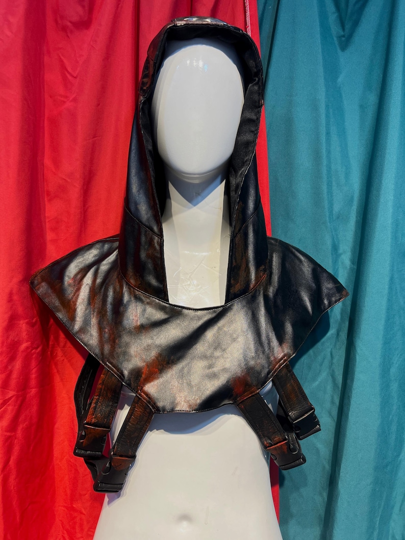 May include: A black faux leather hood and shoulder piece with red accents, displayed on a mannequin. The hood has a pointed top and the shoulder piece has straps with buckles. The background is red and teal.