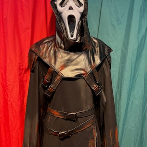 May include: A black Scream costume with a white mask featuring a screaming expression. The costume includes a hooded cloak, shoulder straps, and a belt with buckles. Red paint-like accents are visible on the costume.
