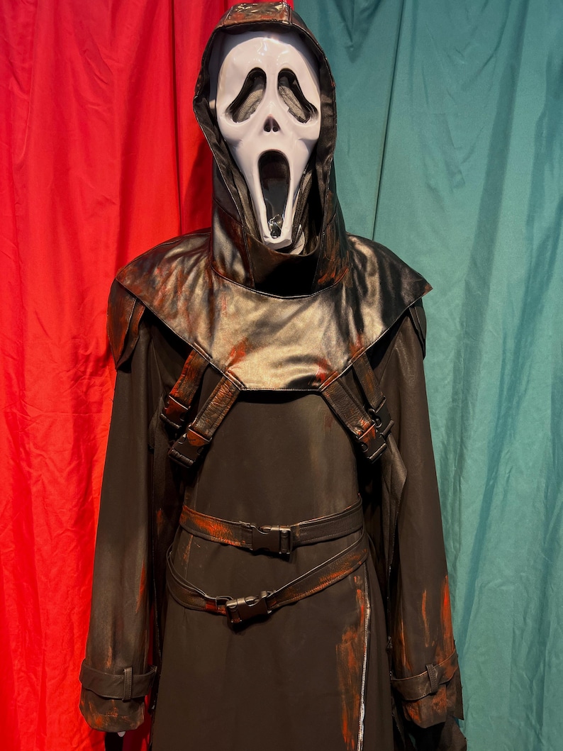 May include: A black Scream costume with a white mask featuring a screaming expression. The costume includes a hooded cloak, shoulder straps, and a belt with buckles. Red paint-like accents are visible on the costume.