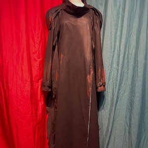 May include: A long, black garment with a high collar and long sleeves. The dress has a distressed appearance with red accents and a jagged hemline. The garment is displayed on a mannequin against a backdrop of red and teal curtains.