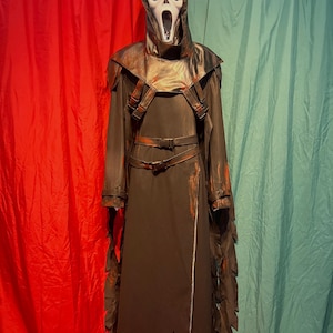 May include: A dark brown Ghostface costume with a white mask, hood, and long robe. The costume features a leather-like chest piece with straps and a belt. The robe has jagged edges and a zipper down the front. The background is red and teal.
