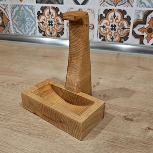 May include: A wooden soap dish and matching soap dispenser. The dispenser is tall and rectangular, with a curved top. The soap dish is rectangular with a curved indentation. Both are made of light-colored wood and sit on a wooden surface.