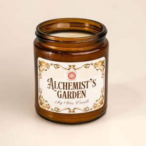 Op de afbeelding: Een bruine glazen potkaars met een wit etiket met de tekst "Alchemist's Garden" in een sierlijk lettertype. Het etiket bevat ook de woorden "Soy Wax Candle". De kaars heeft een wit wasinterieur.