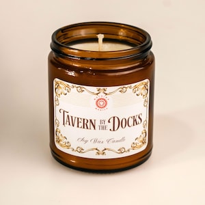 Może przedstawiać: Brązowa świeca w szklanym słoiku z białą etykietą z napisem "Tavern by the Docks" w ozdobnej czcionce. Etykieta zawiera również słowa "Soy Wax Candle". Świeca ma biały knot i brązowy szklany pojemnik.