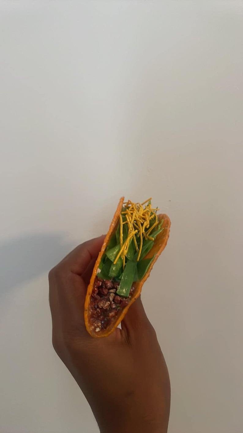 May include: A vibrant orange taco shell filled with green lettuce, dark red beans, and topped with yellow shredded cheese. The taco is held against a plain white background, showcasing the colorful ingredients.