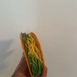 May include: A single taco with a bright orange shell, filled with green lettuce, yellow shredded cheese, and a dark filling. The taco is held against a plain white background.