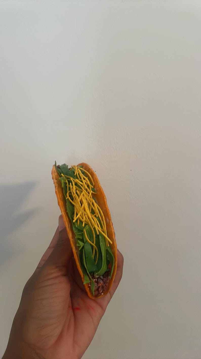 May include: A single taco with a bright orange shell, filled with green lettuce, yellow shredded cheese, and a dark filling. The taco is held against a plain white background.
