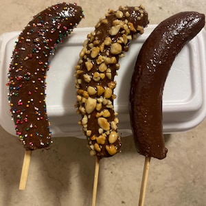 Könnte beinhalten: Drei schokoladenüberzogene Bananen auf Stöcken. Eine Banane ist mit Schokoladenstreuseln bedeckt, eine andere mit gehackten Erdnüssen und die dritte mit Schokolade pur.