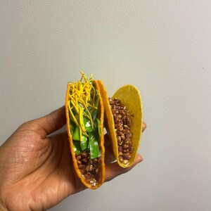 May include: Two yellow hard-shell tacos filled with various ingredients. One taco is filled with brown crumbles, while the other is filled with green and yellow toppings. The tacos are held in a hand.