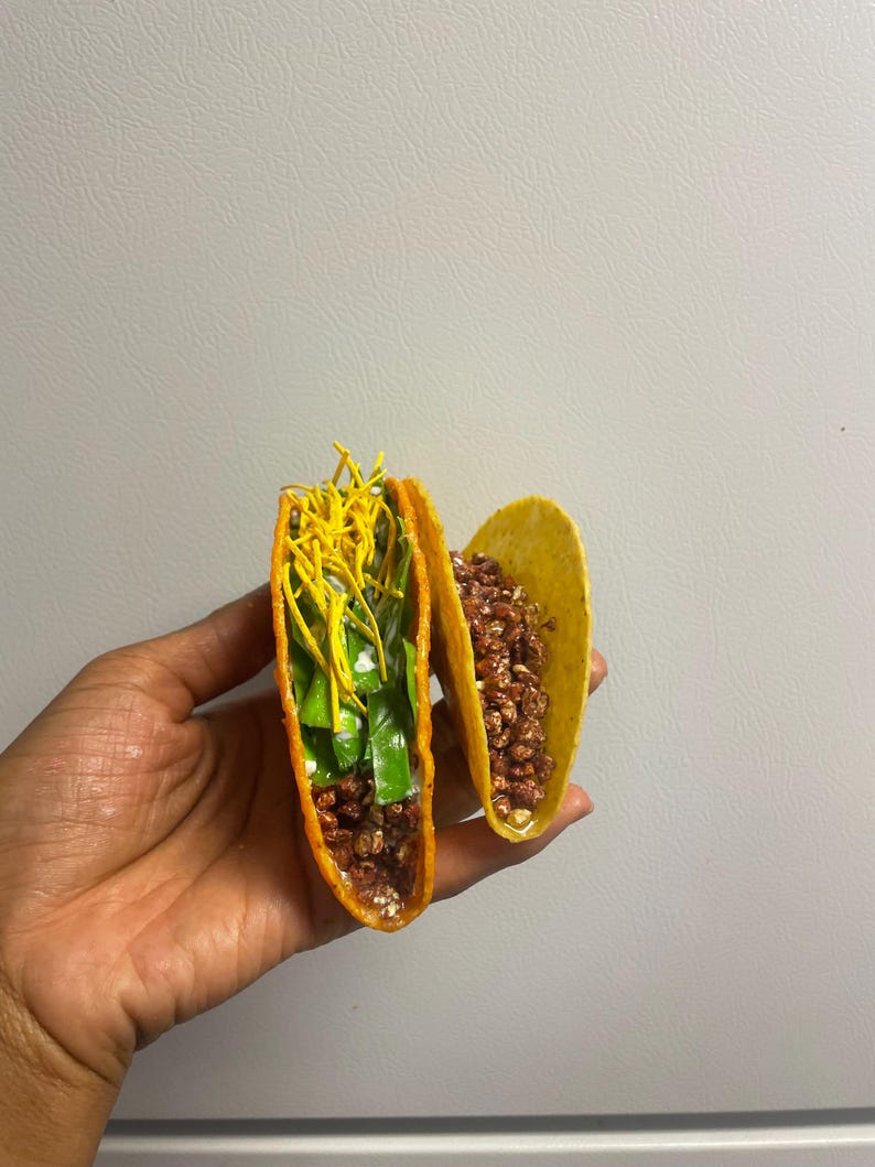 May include: Two yellow hard-shell tacos filled with various ingredients. One taco is filled with brown crumbles, while the other is filled with green and yellow toppings. The tacos are held in a hand.