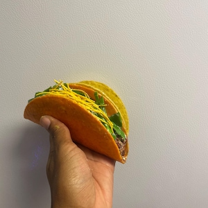 May include: A vibrant orange taco shell filled with green lettuce, yellow shredded cheese, and a dark filling. The taco is held in a hand against a plain white background. The taco appears to be freshly made.