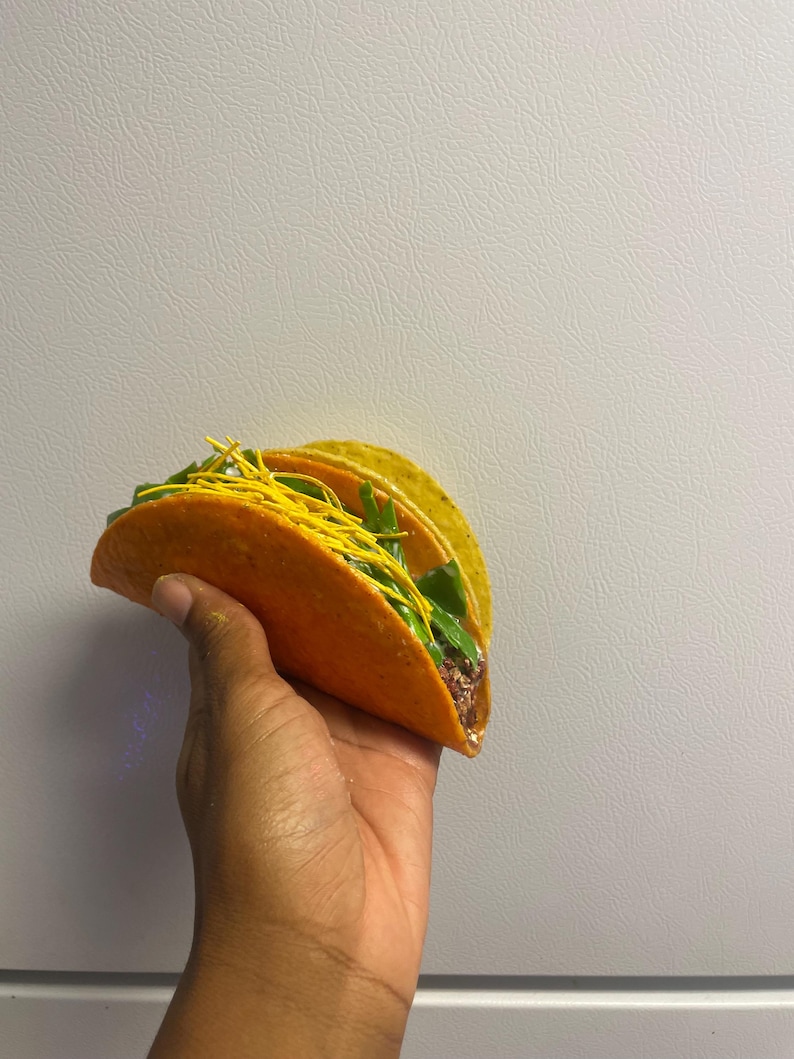 May include: A vibrant orange taco shell filled with green lettuce, yellow shredded cheese, and a dark filling. The taco is held in a hand against a plain white background. The taco appears to be freshly made.