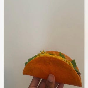 May include: Two orange tacos filled with green lettuce and yellow strands, held by a hand. The tacos have a vibrant orange shell and are filled with fresh ingredients, ready to eat.