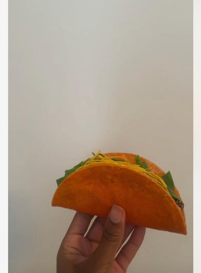 May include: Two orange tacos filled with green lettuce and yellow strands, held by a hand. The tacos have a vibrant orange shell and are filled with fresh ingredients, ready to eat.