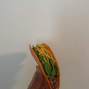 May include: A single taco with a bright orange shell, filled with green leafy vegetables, a reddish-brown filling, and yellow noodle-like toppings. The taco is held against a plain white background.
