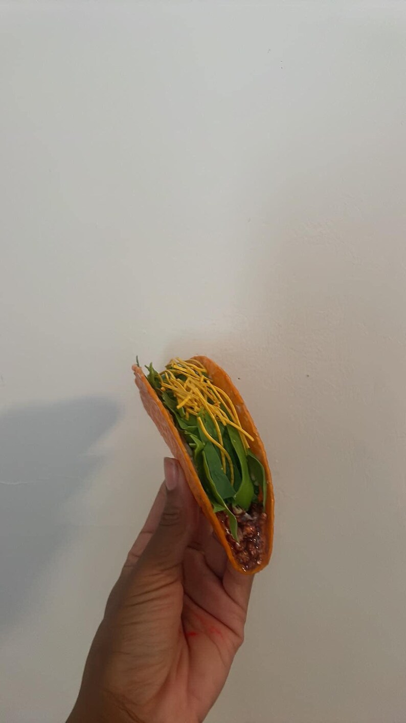 May include: A single taco with a bright orange shell, filled with green leafy vegetables, a reddish-brown filling, and yellow noodle-like toppings. The taco is held against a plain white background.
