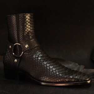 May include: A black leather ankle boot with a snakeskin texture. The boot features a silver-colored metal ring and strap detail around the ankle. The boot has a pointed toe and a small heel.