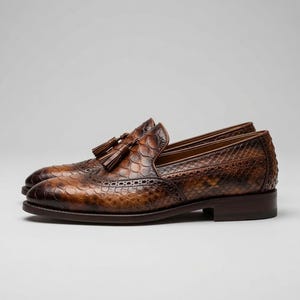 May include: A pair of brown leather loafers featuring a textured snakeskin-like pattern, a decorative tassel on the vamp, and classic brogue detailing along the toe cap. The shoes have a low stacked heel and a polished finish throughout.