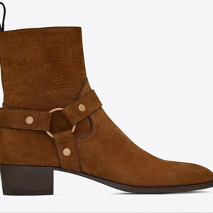 May include: A brown suede ankle boot with a textured crocodile-embossed pattern. The boot features a harness strap across the mid-section secured by a gold-toned metal ring and three matching gold-toned studs. It has a dark block heel and a pull tab.