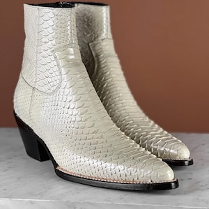 Puede incluir: Botines color crema con un patrón texturizado similar a la piel de serpiente. Los botines tienen punta, tacón negro y cremallera lateral. Los botines están hechos de un material que se asemeja a la piel de serpiente.