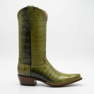 Puede incluir: Una bota alta de estilo vaquero en color verde oliva con un patrón texturizado de relieve de cocodrilo. La bota presenta una punta afilada, un tacón de madera apilado y tiradores en la parte superior de la caña. El acabado va de oliva claro a oscuro.