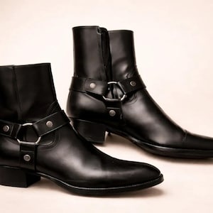 Puede incluir: Un par de botines de cuero negro con punta afilada, tacón de bloque y cierre de cremallera lateral. Cada bota está adornada con una correa decorativa que incluye una anilla de metal plateado y tres remaches metálicos circulares.