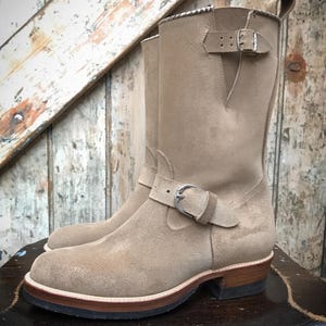 May include: A tan suede mid-calf boot featuring a rounded toe, a stacked wooden heel, and a black rubber outsole. The boot includes a decorative strap across the instep with a silver-toned metal buckle and a matching adjustable strap at the top.