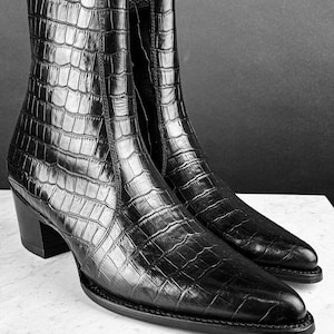 May include: Black leather ankle boots with a crocodile skin effect. The boots feature a pointed toe, a stacked heel, and a zip on the inner side. They are presented on a white surface against a dark backdrop.