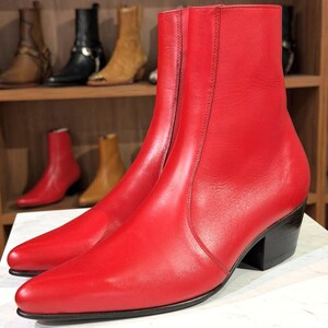 Puede incluir: Botines de cuero rojo brillante con punta afilada y tacón negro. Se ven botas adicionales en varios colores en una estantería de madera en el fondo.