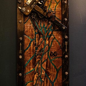 May include: A wooden wall art piece with a dark brown frame and a green and brown resin design. A small, black gun is attached to the resin design.