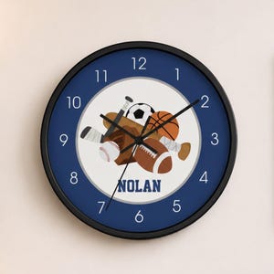 May include: A round wall clock with a navy blue face and black frame. The clock features a white center with sports equipment illustrations and the name "NOLAN". The clock is surrounded by the numbers 1-12.