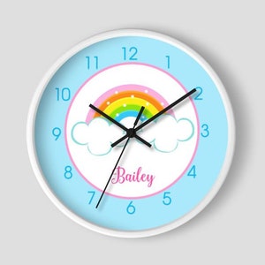 May include: A white clock with a blue background and a rainbow with clouds and stars in the center. The clock face has black hands and pink numbers. The name "Bailey" is written in pink below the rainbow.