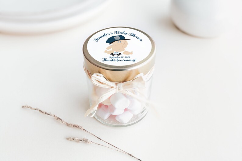 May include: A small glass jar with a gold lid and a white ribbon tied around it. The jar is filled with small white and pink marshmallows. The lid has a round sticker with a cartoon baby wearing a police hat and holding a toy police car. The sticker says "Jennifer's Baby Shower" and "September 22, 2019" and "Thanks for coming!"