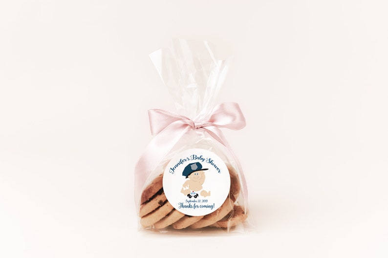 May include: A clear plastic bag filled with cookies tied with a pink ribbon. A round sticker with a cartoon baby wearing a police hat and the text "Jennifer's Baby Shower September 27, 2020 Thanks for coming!" is attached to the bag.