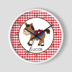 May include: White clock with red and white gingham pattern around the edge. The center of the clock features a cartoon brown horse with a blue and red saddle. The horse is wearing a red star. The clock face has black numbers and hands. The name "Lucas" is printed below the horse.