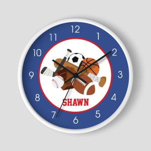 May include: A white round clock with blue and red accents. The clock face features a white background with a red border and a blue inner circle. The inner circle features a variety of sports equipment, including a baseball, basketball, football, soccer ball, and bats. The name "SHAWN" is printed in red letters below the sports equipment. The numbers 1 through 12 are printed in white around the outer edge of the clock face.