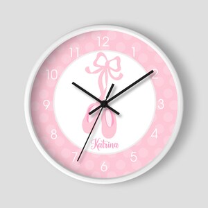 May include: A white clock with pink polka dots and a pink ballerina shoe design. The clock face has black hands and black numbers. The name "Katrina" is written in black cursive below the ballerina shoes.