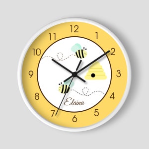 May include: A yellow and white round wall clock with black hands and bee illustrations. The clock face has black numbers 1 through 12 and the name "Elaina" written in brown cursive.