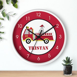 May include: Red and white round wall clock with a fire truck and a dalmatian dog on it. The clock has black hands and white numbers. The name "TRISTAN" is printed below the fire truck.