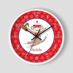 May include: A white clock with red polka dots and a cartoon monkey wearing a knitted hat and holding a heart. The clock face has black numbers and hands. The monkey's name is Teddy.