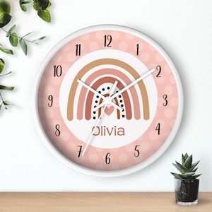 May include: A pink and white wall clock with a rainbow design and the name "Olivia" printed on the face. The clock has black polka dots around the edge and a white frame.