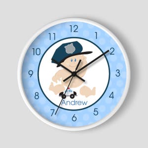 May include: A white clock with blue polka dots and a cartoon baby wearing a police hat and holding a toy car. The clock face has black numbers and hands. The baby's name, "Andrew", is printed below the car.