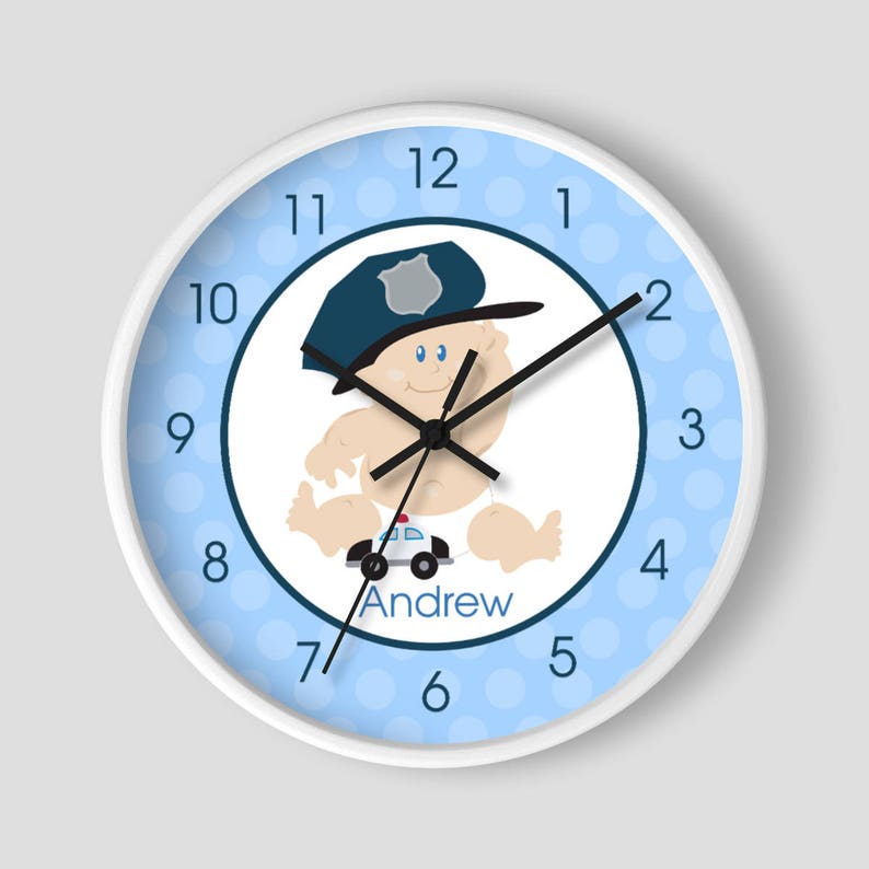 May include: A white clock with blue polka dots and a cartoon baby wearing a police hat and holding a toy car. The clock face has black numbers and hands. The baby's name, "Andrew", is printed below the car.