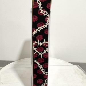 May include: A decorative bookmark with a black background featuring red roses and a white thorn design. The bookmark is held in a clear plastic holder, showcasing the floral pattern.