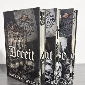 May include: Three hardcover books with black and white covers featuring gothic illustrations of skulls, ornate architectural arches, and wax seals with the letter B. The first book is titled Deceit by Katelyn Taylor, while the others show partial text.