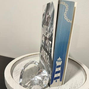 May include: A book titled "Does It Hurt?" with a black and white image of a lighthouse on the cover. The book is standing upright on a white wooden tray with a clear crystal heart.