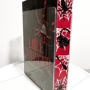 K&ouml;nnte beinhalten: Ein Buchdisplay mit dem Buch "Little Stranger" von Leigh Rivers. Das Display hat eine transparente Vorderseite und ein rotes und wei&szlig;es Spinnennetz-Design an der Seite. Das Buchcover zeigt eine gro&szlig;e rote Spinne.
