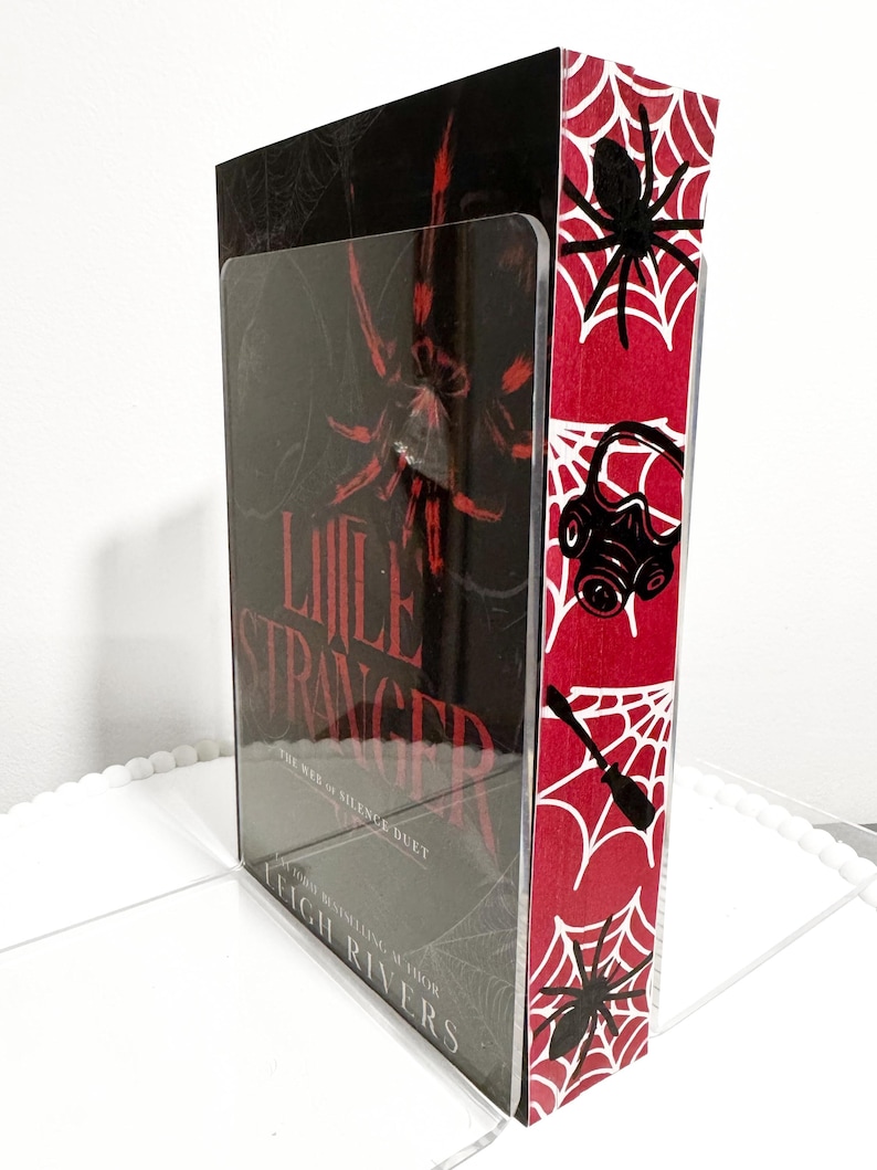 K&ouml;nnte beinhalten: Ein Buchdisplay mit dem Buch "Little Stranger" von Leigh Rivers. Das Display hat eine transparente Vorderseite und ein rotes und wei&szlig;es Spinnennetz-Design an der Seite. Das Buchcover zeigt eine gro&szlig;e rote Spinne.
