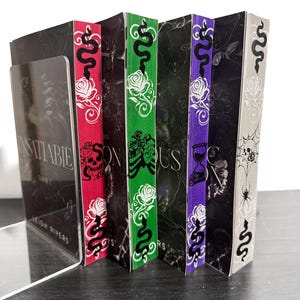 May include: A collection of fantasy books with unique spine designs. The spines are red, green, purple, and gray, each featuring a snake, rose, and other gothic-style illustrations. The book covers have a dark, marble-like texture.