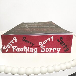 K&ouml;nnte beinhalten: Zwei B&uuml;cher auf einer wei&szlig;en Oberfl&auml;che gestapelt. Das obere Buch hat einen dunklen Einband. Das untere Buch hat einen roten Einband mit den Worten "Sorry" und "Fucking Sorry" in wei&szlig;er Schrift. Die B&uuml;cher stehen auf einem wei&szlig;en Perltablett.